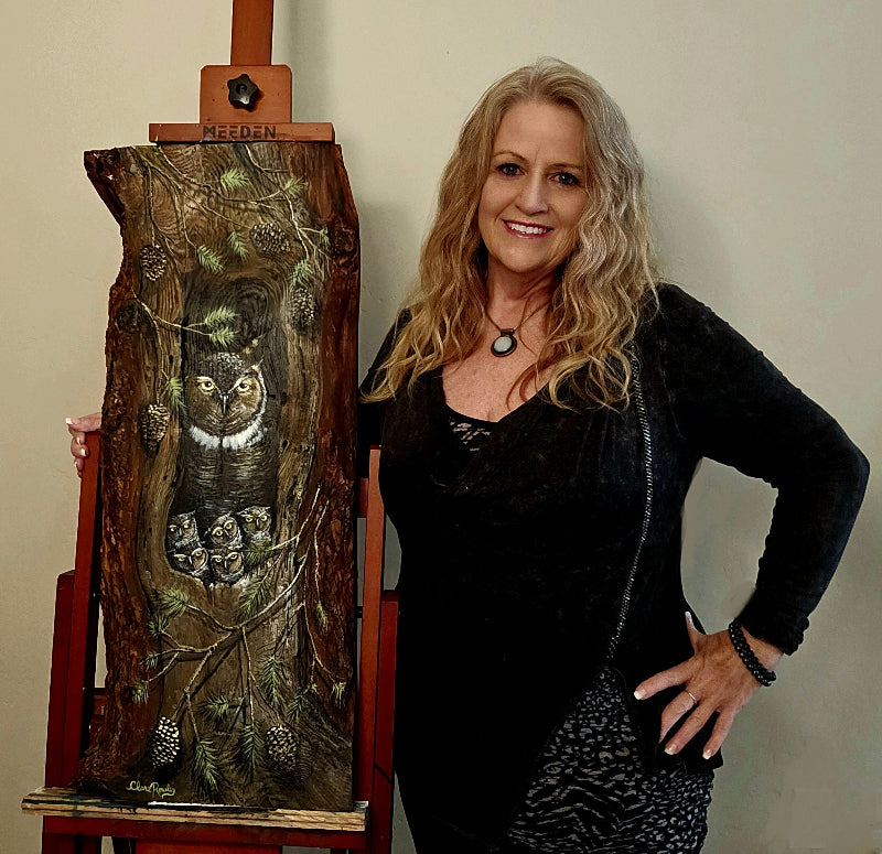 cream color background with Clare Rowley standing in front of it wearing a black and grey outfit and her hand is wrapped around her  painting on wood - the painting is a great horned owl family painted on Durango Colorado Pine 